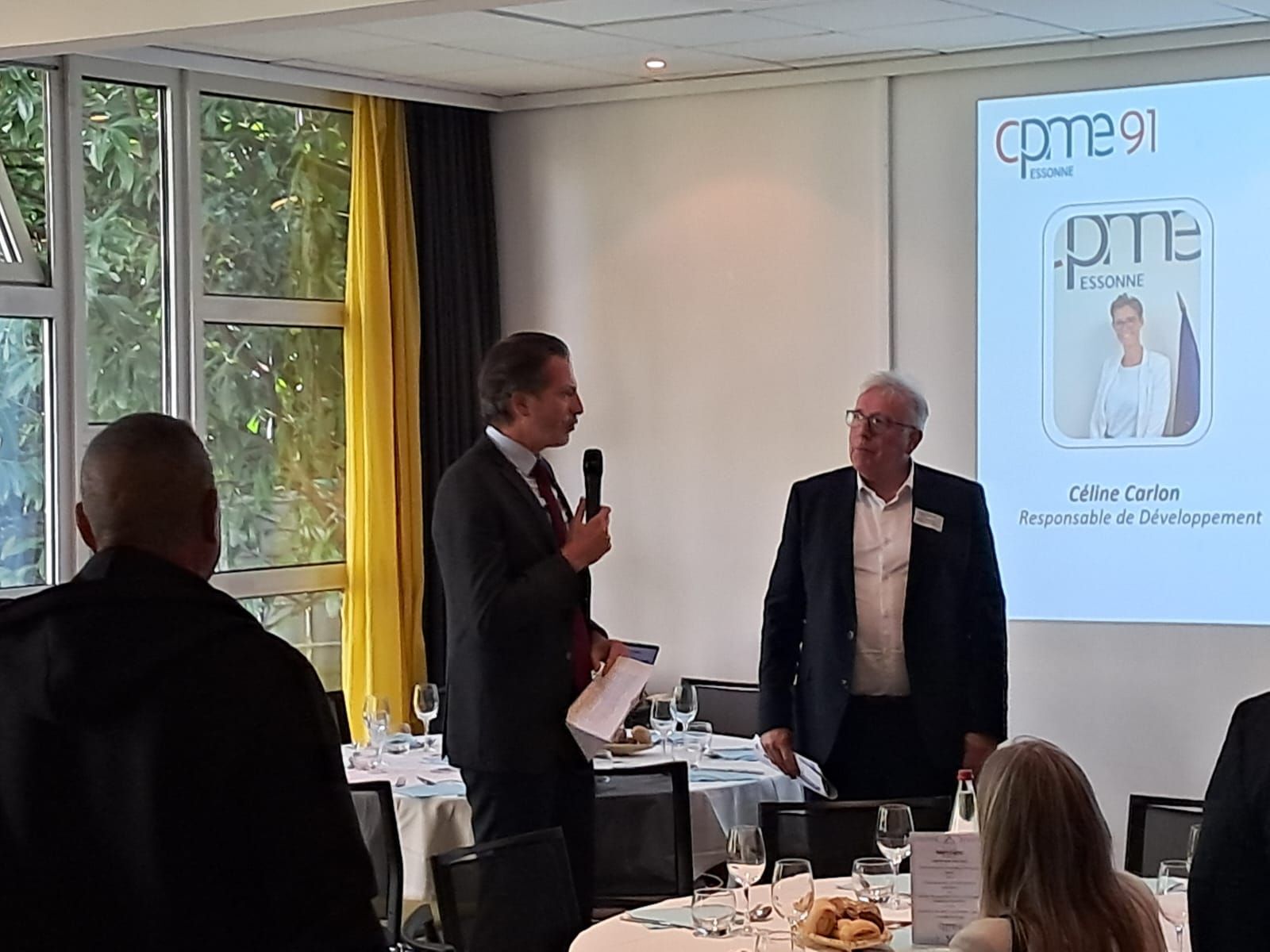 Dejeuner CPME 91 avec présentation sur l'écran en arrière plan, 1 homme avec micro et 1 autre homme debout à coté. L'audience est autour de table de déjeuner.