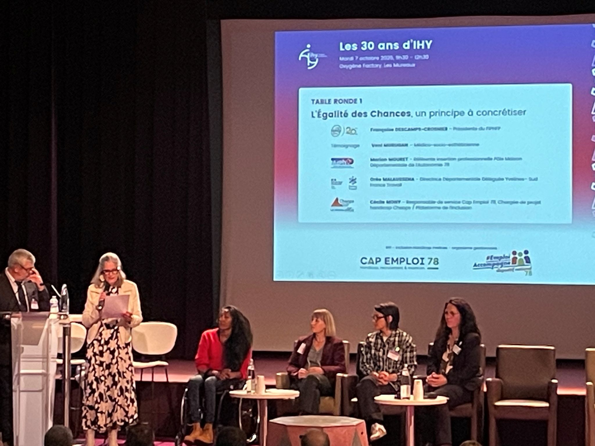 Scène des 30 de CAP Emploi 78 - IHY au Campus Oxygène Factory – Les Mureaux. 1 homme debout au pupitre, 1 femme debout à coté lisant son texte, 4 femmes assises, 1 présentation projetée en arrière plan sur l'écran